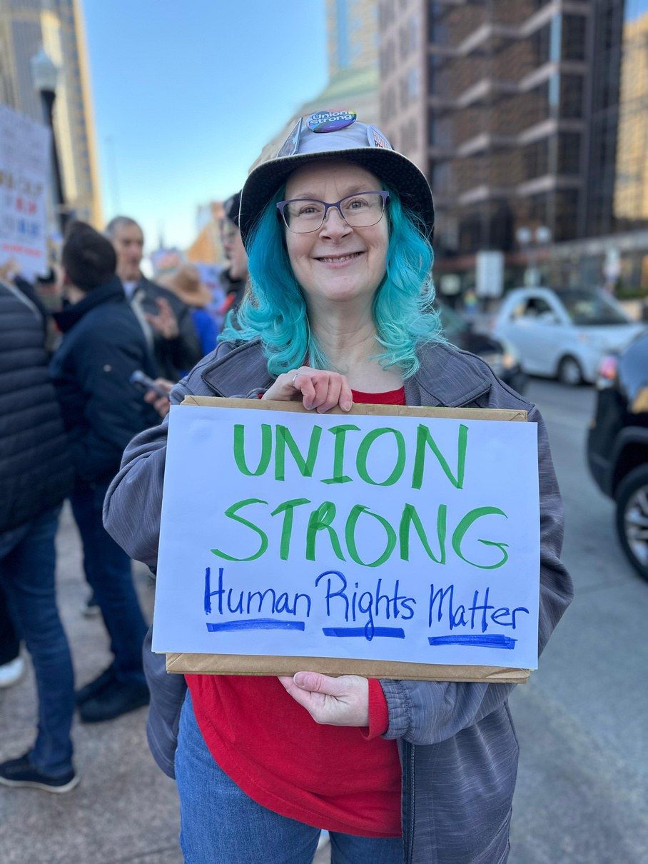 A smiling demonstrator in a Union Strong bucket hat holds a hand-lettered sign reading UNION STRONG — Human Rights Matter at a street rally, with other marchers behind her.
